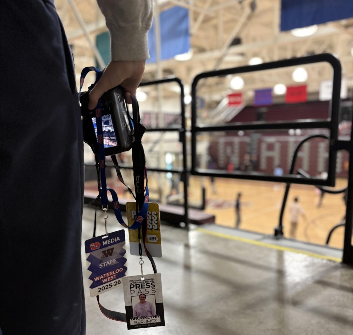 Mackenzie Johnson holding a camera and multiple press passes while in the West High school main gym.